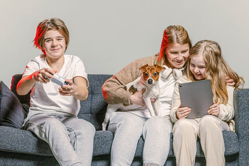 Äiti, lapsi ja koira istuvat sohvalla ja käyttävät älylaitteita.