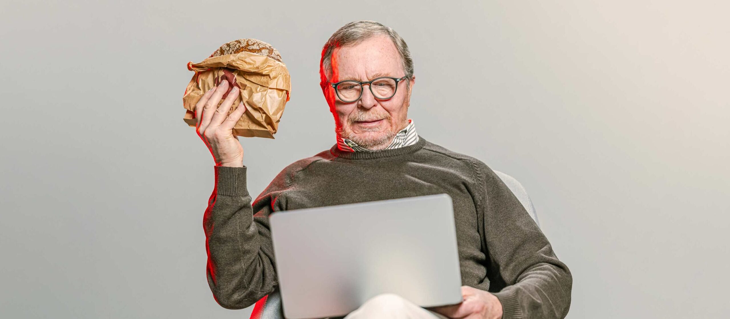 Mies istuu kannettava tietokone sylissään ja pitelee kädessään ruisleipää.